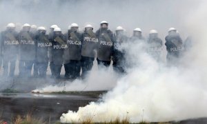 Policjanci idą w rzędzie z tarczami i przechodzą przez dym.