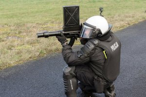 Policjant klęczy na jedno kolano. W ręku trzyma wyrzutnik granatów łzawiących.