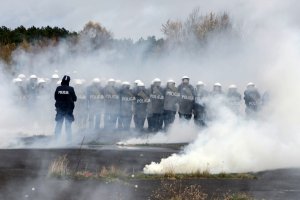 Policjanci idą w rzędzie z tarczami i przechodzą przez dym.