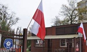 Główna brama wjazdowa do słupskiej Szkoły Policji. Na słupach trzymających skrzydła bramy zawieszone biało-czerwone flagi. Ujęcie z lewej strony.