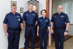 Czterej stojący policjanci na tle białej ściany i stojącego za nimi dużego zegara.