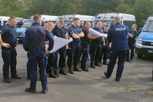 Stojący policjanci, w tle zaparkowane radiowozy.